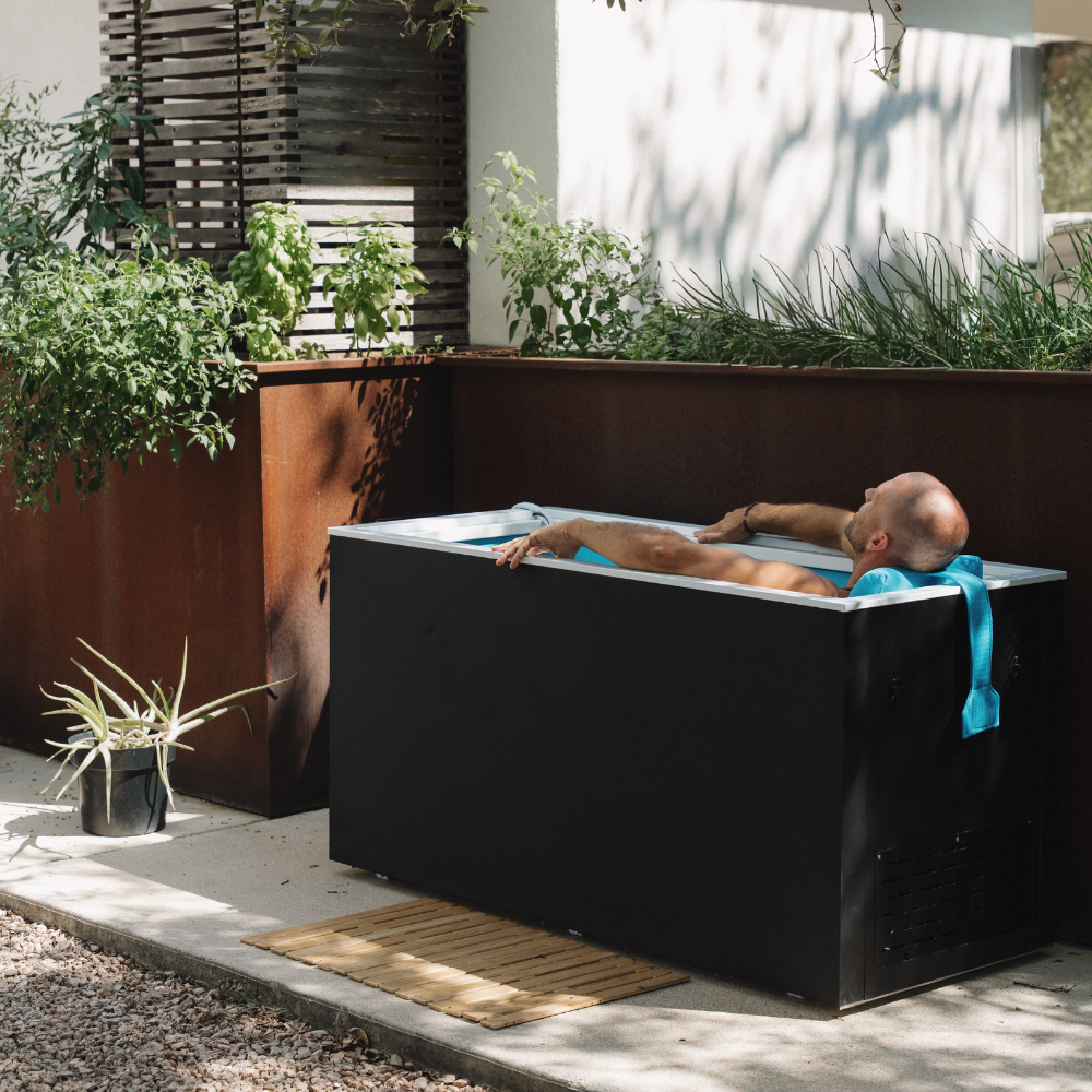 A man lying in a Black matte Plunge Zero SoftTop cold plunge tub with plants and a wooden deck in the background
