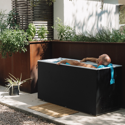 A man lying in a Black matte Plunge Zero SoftTop cold plunge tub with plants and a wooden deck in the background
