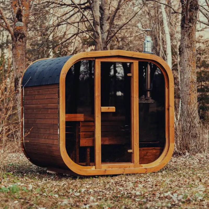 Fully assembled Mande Spa USA MW20 outdoor sauna with wood-burning heater, tempered glass door and windows, Bituminous roof tiles, and metal bands in a peaceful forest setting.