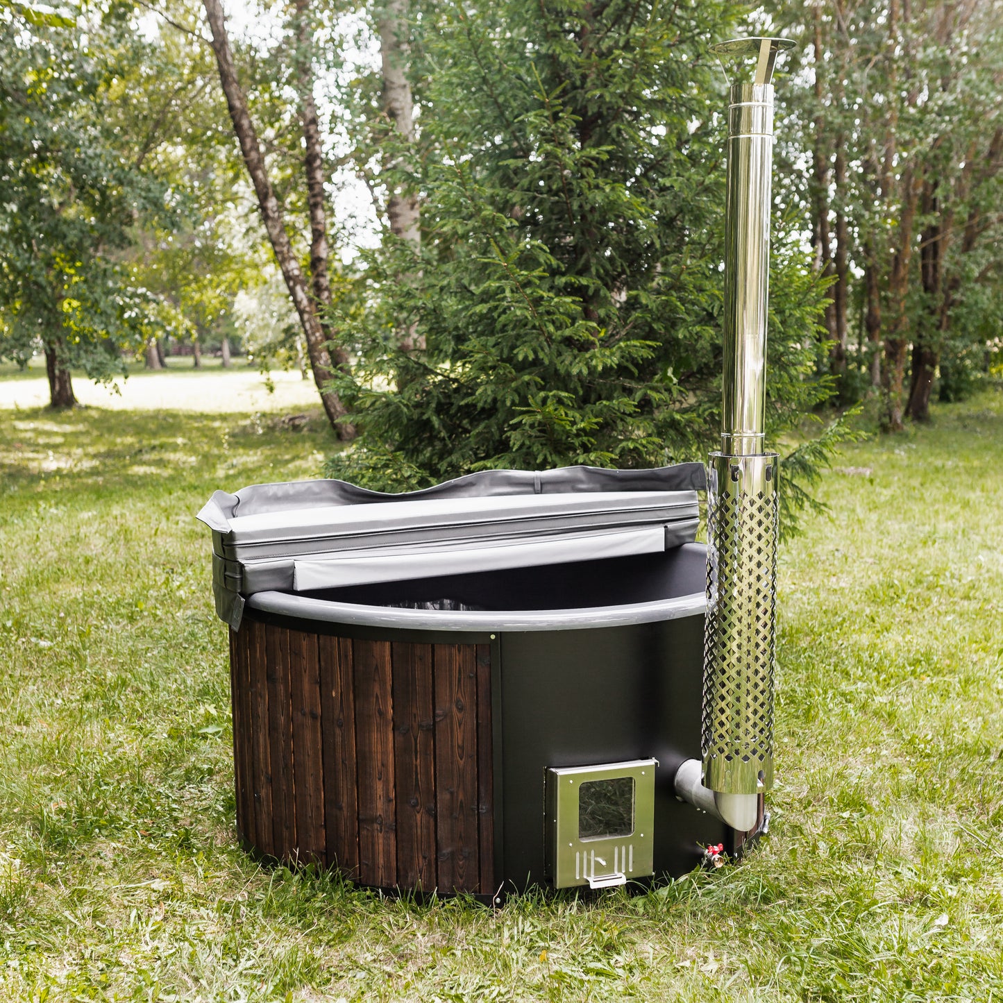 Outdoor hot tub with wooden exterior and metal chimney in a grassy area with trees.