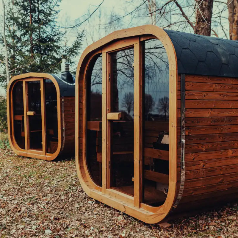 Two fully assembled Mande Spa USA MW16 outdoor saunas with tempered glass doors and windows, showing a wood-burning heater model on the left and an electric heater model on the right.